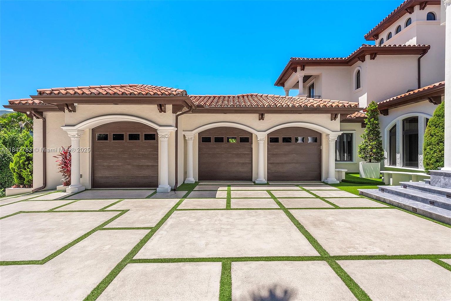 This image showcases a Mediterranean-style home featuring a three-car garage with arched, wood-toned doors and decorative columns. The driveway is constructed of large concrete pavers separated by strips of green grass, creating a modern, geometric aesthetic. The exterior walls are finished in a light stucco, complemented by a classic terracotta tile roof.