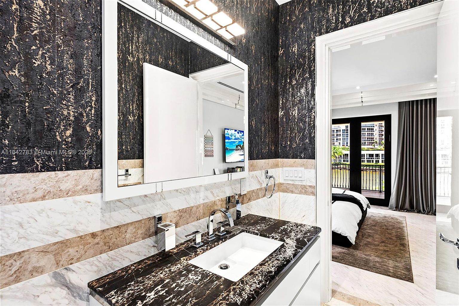 This modern bathroom features a striking vanity with a dark, patterned stone countertop and a white rectangular sink. The walls are adorned with a bold, textured black and gold wallpaper above a horizontal band of light-toned stone tile. A large, rectangular mirror is centered above the vanity, and a doorway provides a clear view into an adjacent bedroom with a bed and balcony access.