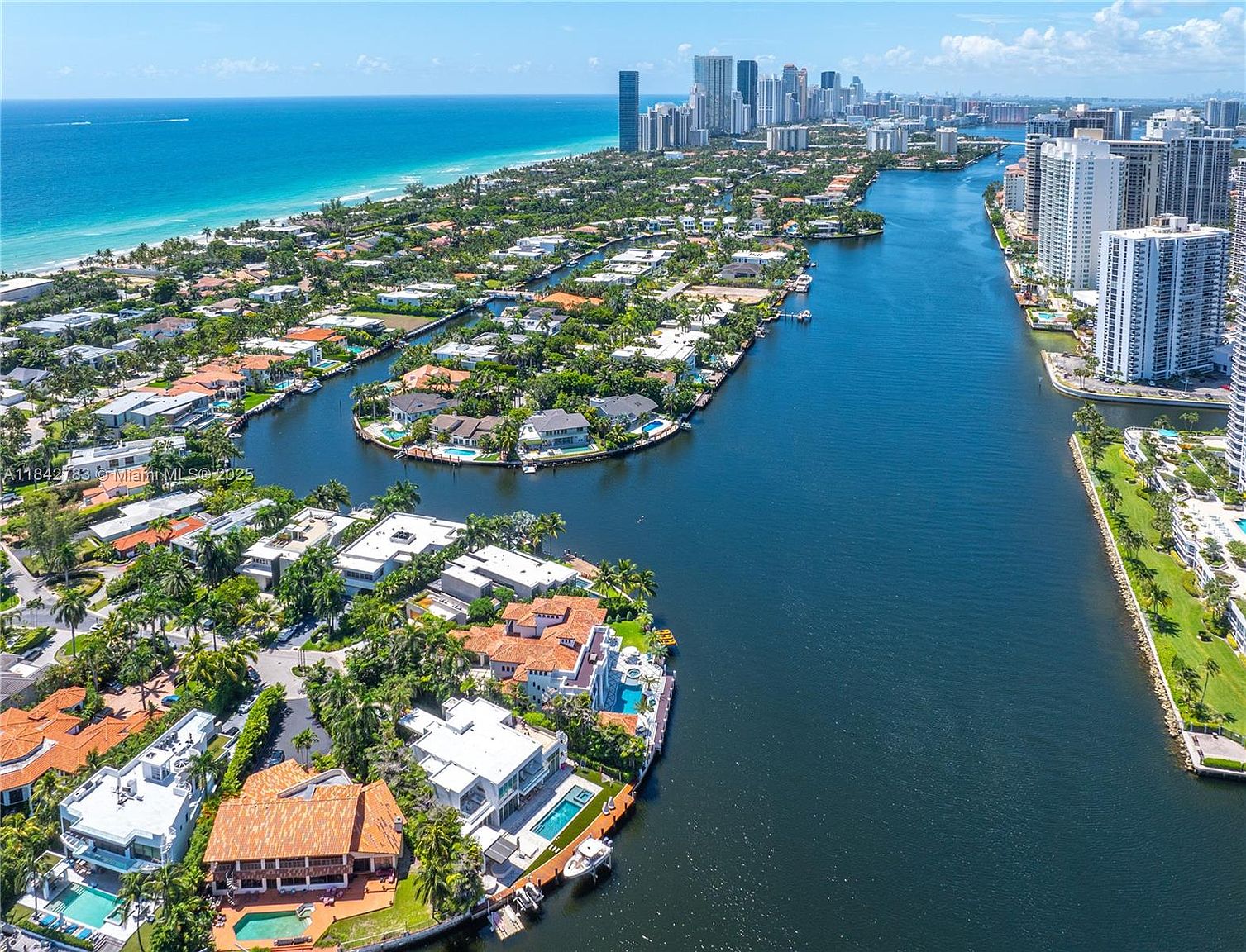 This high-altitude aerial view captures a luxurious waterfront residential neighborhood in Florida, characterized by winding canals and lush tropical landscaping. The perspective showcases numerous multi-story homes with private docks and swimming pools, set against the backdrop of the Atlantic Ocean and a distant skyline of high-rise buildings. The scene emphasizes the prime coastal location and the serene, upscale lifestyle offered by this waterfront community.