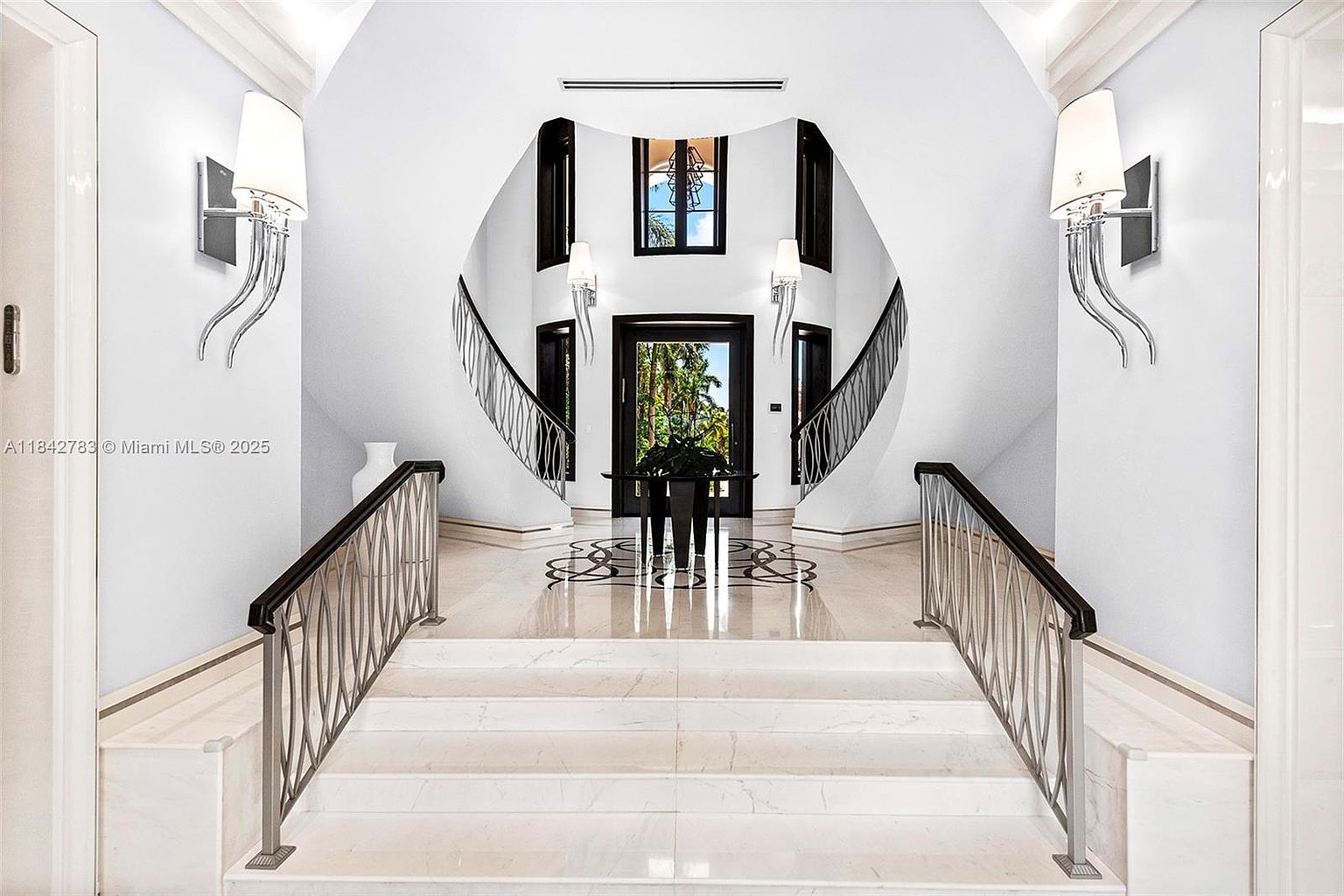 This grand foyer features a symmetrical staircase design with elegant metal railings leading up to a landing. The space is finished in a bright, neutral palette with polished stone flooring and decorative wall sconces that provide soft illumination. A central console table sits at the base of the stairs, framing a view through the front doorway toward the lush greenery outside.