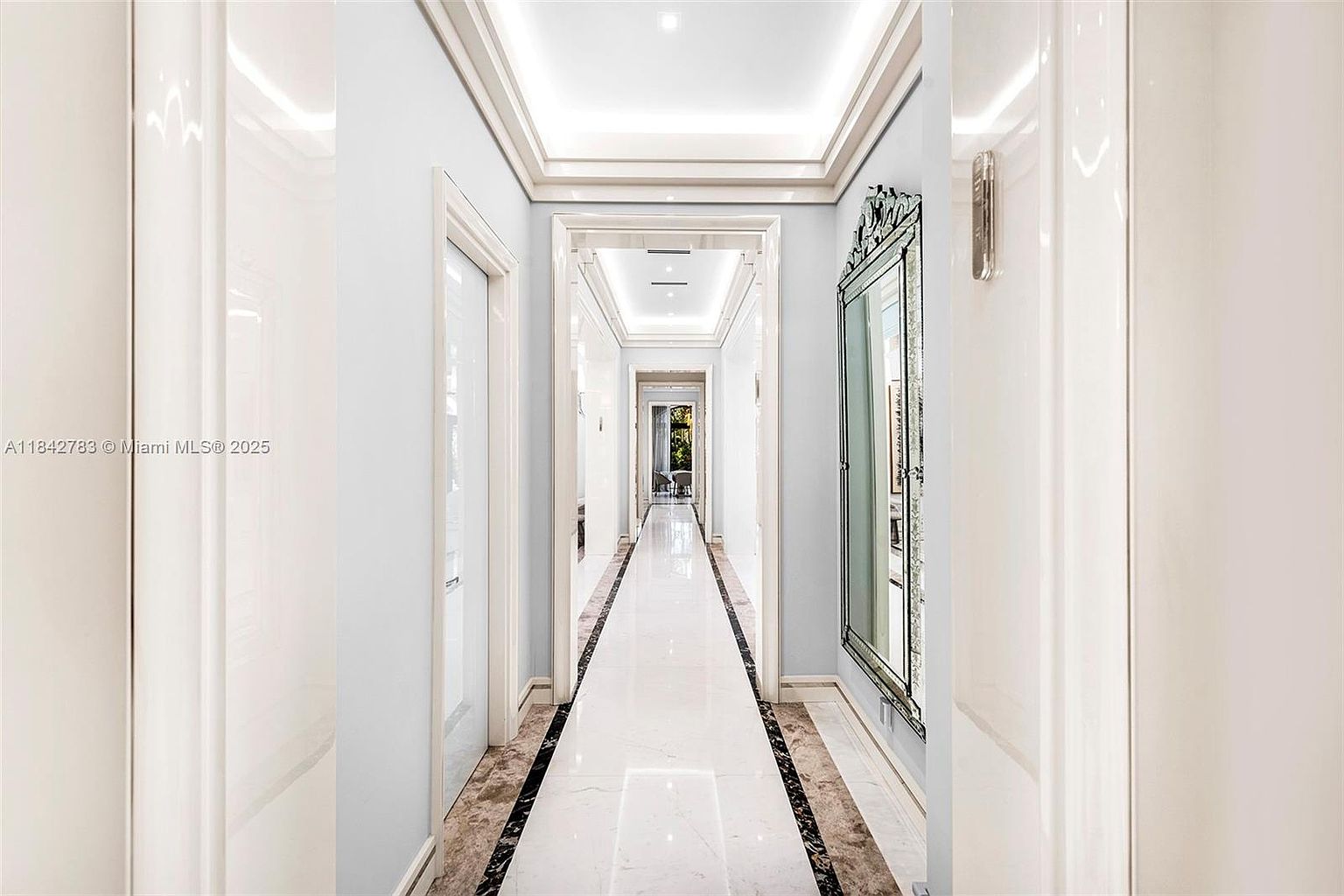 This elegant, long hallway features a polished light-colored stone floor with a dark stone border, creating a sophisticated linear perspective. The walls are painted a soft, pale blue, complemented by a large, ornate mirror on the right and recessed ceiling lighting that adds depth to the corridor. The view extends through multiple arched doorways, emphasizing the grand scale and symmetrical design of the home's interior architecture.