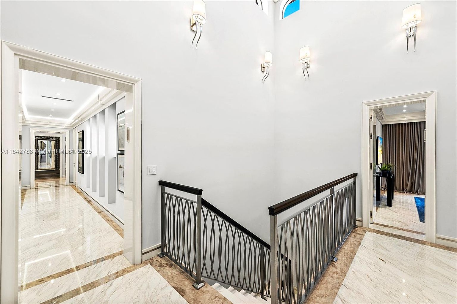 This interior view captures a grand landing area featuring a staircase with a decorative metal railing and a dark wood handrail. The space is characterized by high ceilings, light-colored walls adorned with wall sconces, and polished stone flooring with a subtle veining pattern. To the left, a long hallway leads toward a mirror, while a doorway on the right opens into a room with draped curtains, creating an elegant and spacious atmosphere.