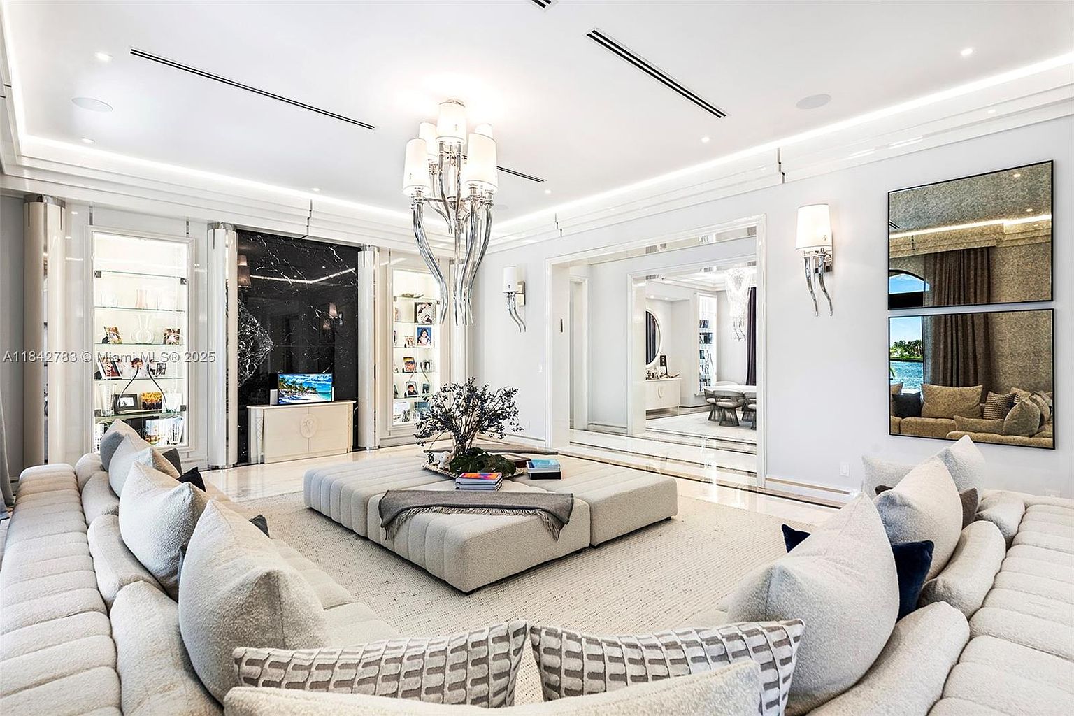 This expansive, ultra-modern living room features a large, plush sectional sofa arranged around a central, oversized square ottoman. The space is defined by a sophisticated monochromatic palette, highlighted by a striking black stone feature wall, recessed ceiling lighting, and a dramatic sculptural chandelier. A wide opening leads to an adjacent room, while mirrored wall panels reflect the bright, airy atmosphere and provide a sense of depth.