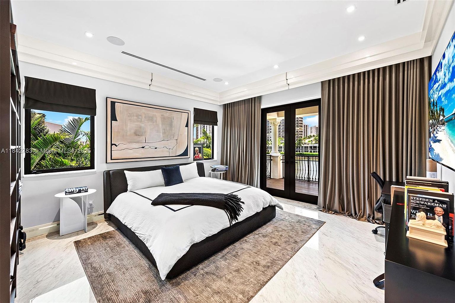 This spacious primary bedroom features a contemporary design with a large bed centered against a wall adorned with a large abstract painting. The room includes light-colored stone flooring, a dark area rug, and tall windows with dark shades, alongside a set of glass doors that open to a private balcony. A desk area is positioned to the right, and the overall aesthetic is sophisticated and minimalist with a neutral color palette.