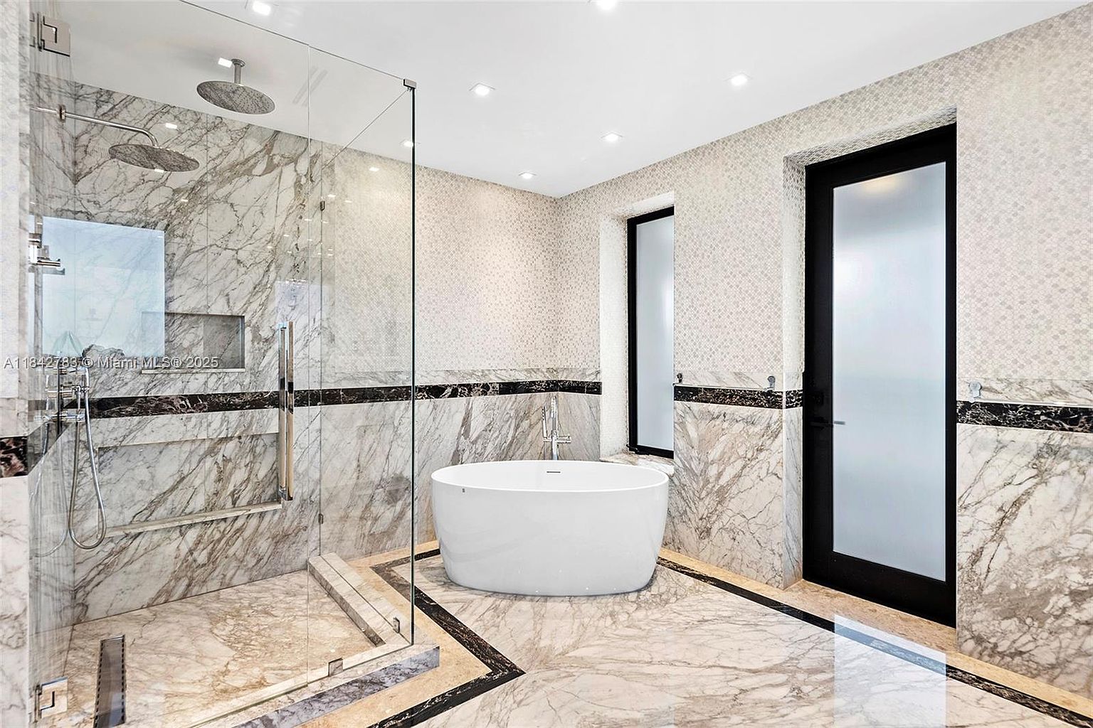This luxurious primary bathroom features a large walk-in glass shower and a freestanding soaking tub as its central focal points. The space is finished with extensive stone wall cladding and patterned tile accents, complemented by a black-framed glass door. The perspective is wide-angle, capturing the high-end, spa-like atmosphere of the room.
