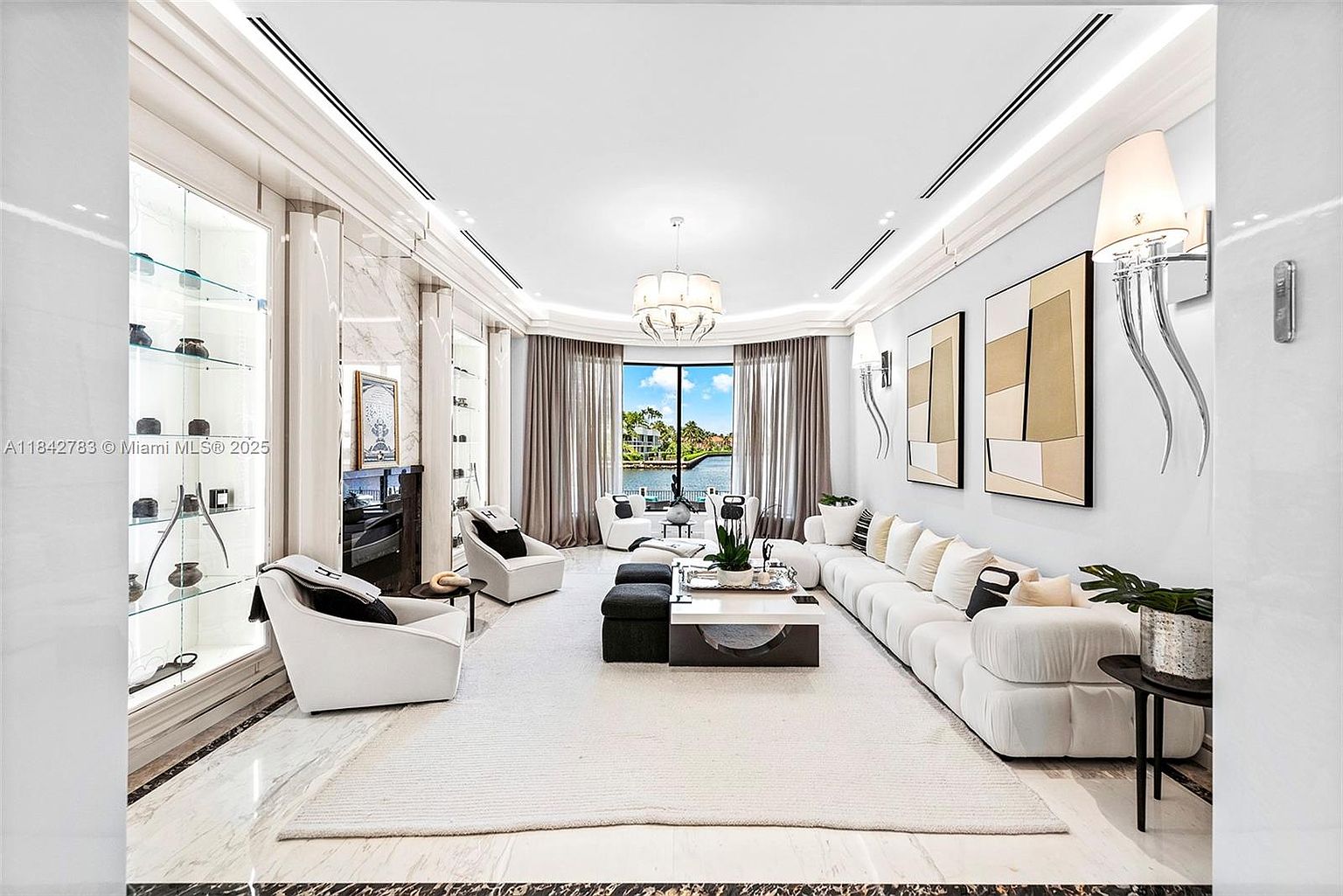 This sophisticated living room features a bright, open-concept design with a large white sectional sofa and matching armchairs arranged around a central coffee table. The space is anchored by a large area rug on a light-toned stone floor, with a tall, stone-clad fireplace surround and a built-in glass display cabinet on the left. Large windows with elegant drapery frame a scenic water view, while modern wall sconces and a central chandelier provide refined lighting.