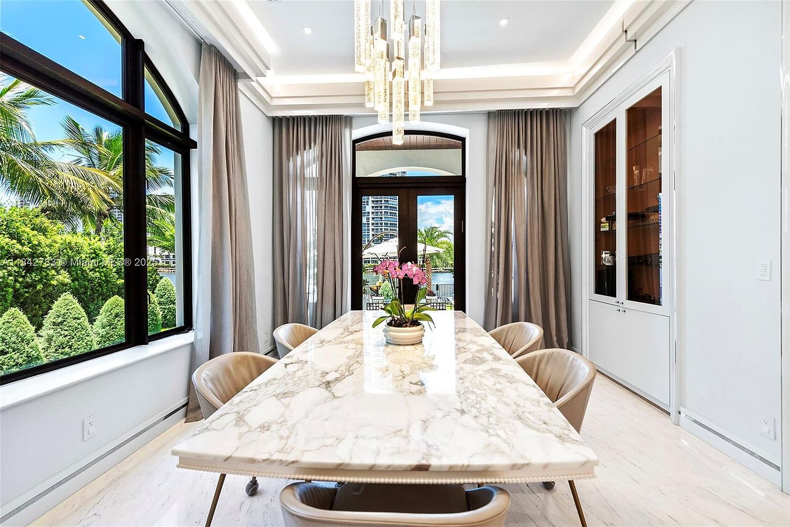 This elegant dining room features a long, light-toned stone table centered beneath a modern, multi-tiered crystal chandelier. Large windows and glass-paned doors provide abundant natural light and views of the lush greenery and waterfront outside. The space is framed by floor-to-ceiling curtains and includes a built-in glass-front display cabinet, creating a sophisticated and airy atmosphere.