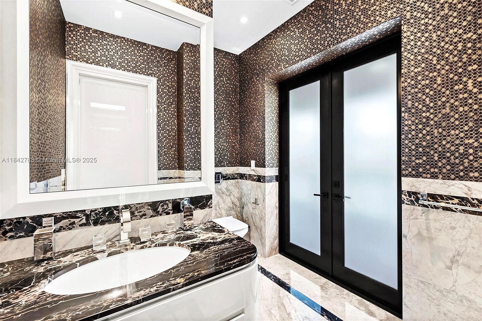 This sophisticated powder room features a striking vanity with a dark, heavily veined stone countertop and a white oval basin. The walls are adorned with a textured, metallic-toned geometric wallpaper, complemented by a large mirror and a double-door entry with frosted glass panels. The space is finished with light-toned stone flooring and a marble-patterned wainscoting, creating a luxurious and high-contrast aesthetic.