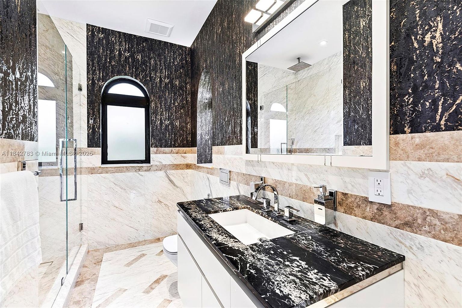 This modern bathroom features a striking contrast between dark, textured wall surfaces and light-toned stone wall tiles. A sleek vanity with a dark, patterned stone countertop and a rectangular undermount sink sits beneath a large, framed mirror. To the left, a glass-enclosed shower area adds to the contemporary aesthetic, while an arched window provides natural light.