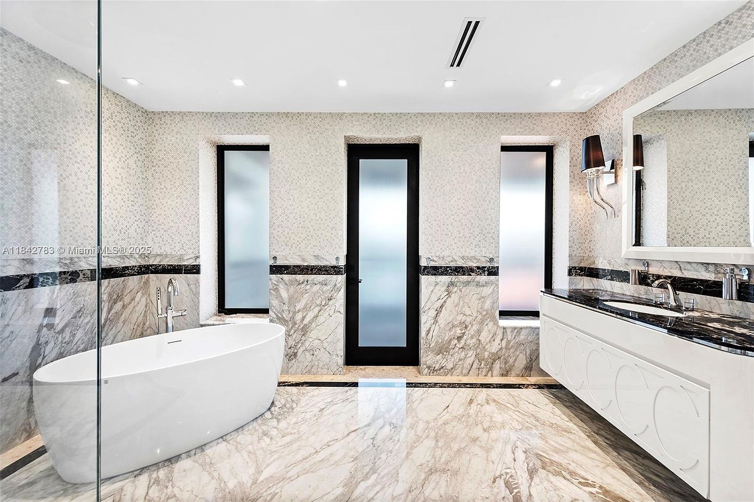 This luxurious primary bathroom features a sleek, freestanding white soaking tub positioned against a backdrop of dramatic, veined stone wall cladding. A long vanity with a dark stone countertop and dual sinks provides ample space, complemented by a large, wide mirror. The room is accented by a central door with frosted glass panels and tall, narrow windows that allow for soft, diffused natural light.