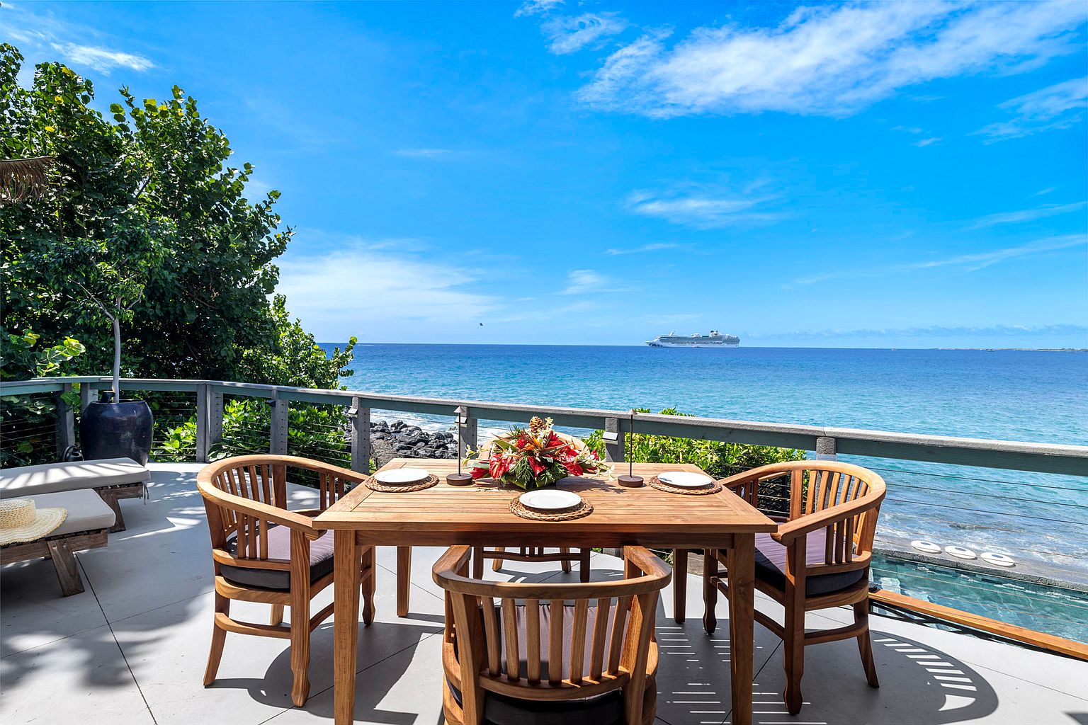 This image showcases a beautifully arranged outdoor dining area on a patio or deck, offering stunning ocean views. The wooden table is set for a meal, complete with a floral centerpiece, and is surrounded by matching chairs with comfortable cushions. The scene evokes a sense of luxury and relaxation, perfect for enjoying meals with a breathtaking backdrop.