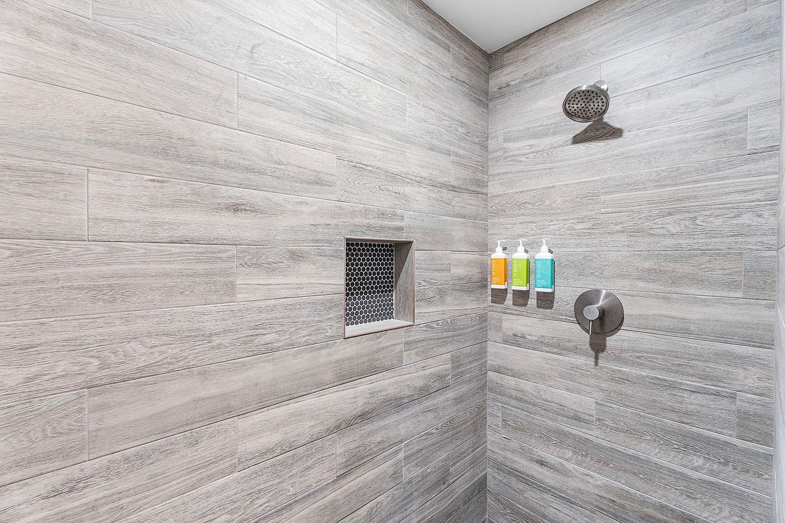 This is a well-lit bathroom featuring a modern shower with gray wood-look tile walls. The shower includes a recessed niche with dark mosaic tile, a rainfall showerhead, and a sleek, brushed metal faucet. Three bottles of bath products are neatly arranged on the wall, adding a touch of color.