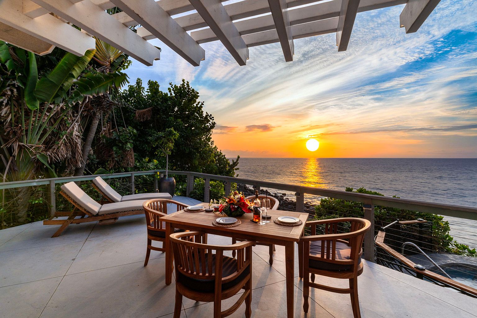 This image showcases a stunning outdoor patio or deck area, perfect for enjoying ocean views and sunsets. The space is furnished with a wooden dining table set for four, complemented by comfortable lounge chairs. A pergola provides partial shade, while the ocean and sunset create a breathtaking backdrop, enhancing the property's appeal for relaxation and entertainment.