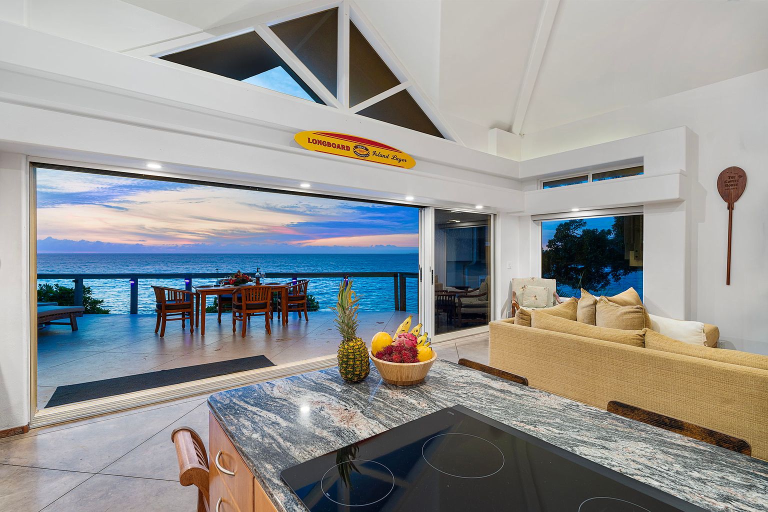 This interior shot showcases a modern kitchen that seamlessly blends indoor and outdoor living. The kitchen features a granite countertop island with a cooktop, while large sliding glass doors open to a patio with ocean views. The open floor plan connects the kitchen to a living area with a comfortable sofa, creating an inviting and luxurious atmosphere.