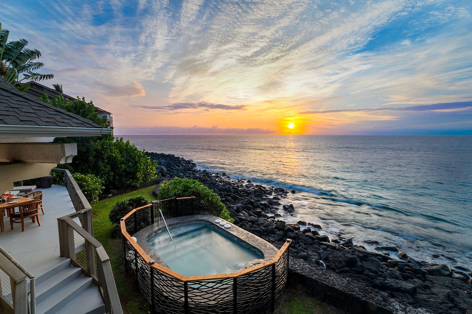 This image showcases a luxurious outdoor spa area at sunset. The spa is situated near the ocean, with a rocky shoreline visible in the background. The setting includes a well-maintained patio area with outdoor furniture, suggesting a relaxing and upscale living environment.