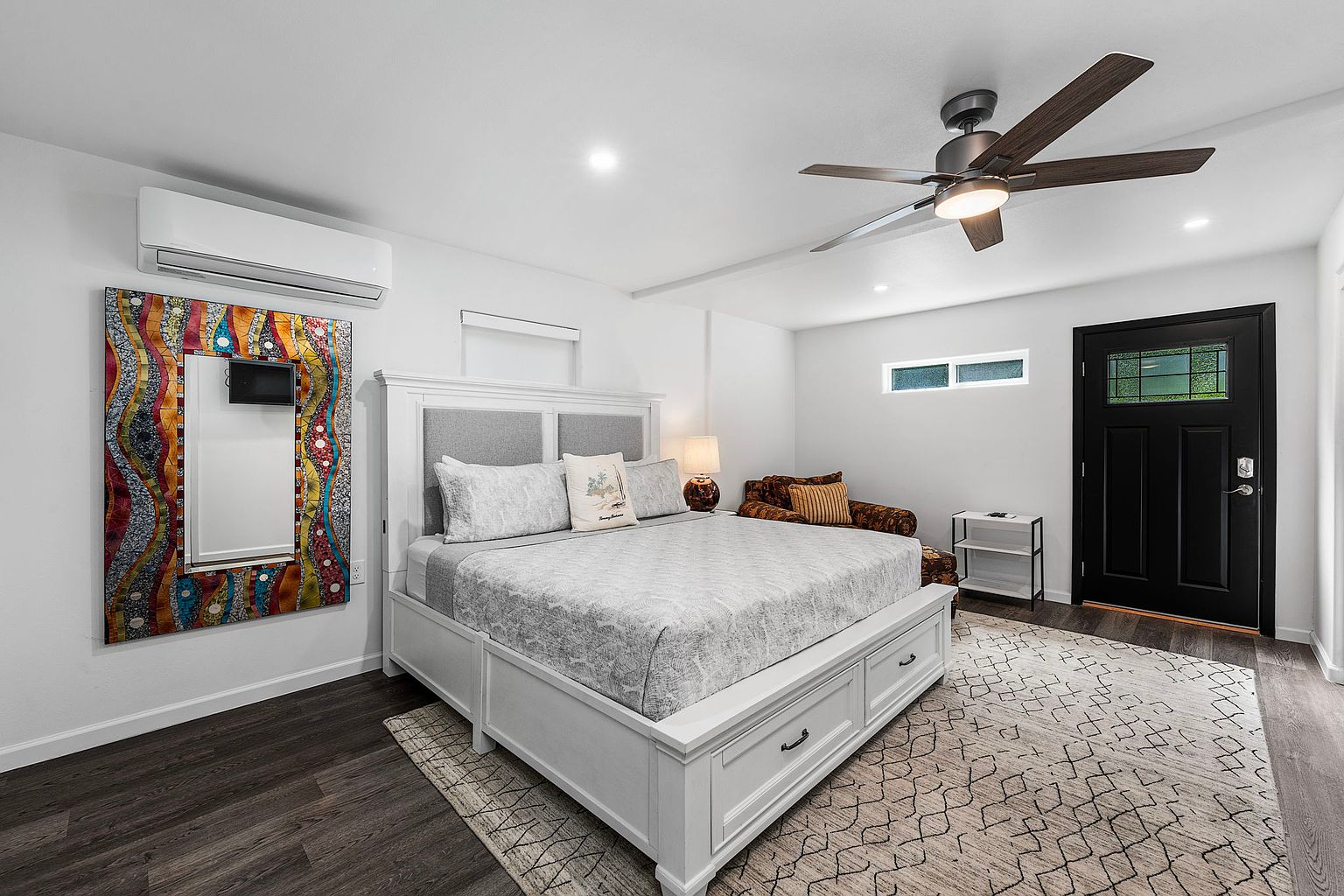 This is a well-lit primary bedroom featuring a large bed with a white frame and gray headboard, complemented by neutral-toned bedding and decorative pillows. A colorful mirror adds a unique touch to the room, while a ceiling fan provides air circulation. The room is finished with a patterned area rug and dark wood flooring, creating a cozy and inviting atmosphere.