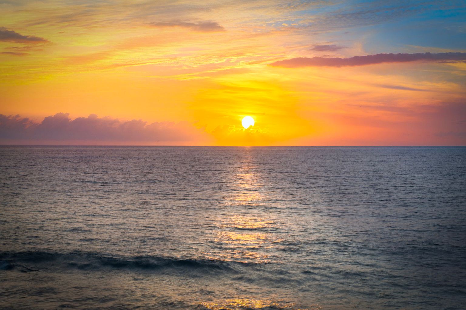 This image showcases a breathtaking seascape at sunset, featuring a vibrant orange and yellow sky reflecting on the calm ocean waters. The sun is positioned near the horizon, creating a stunning focal point with a shimmering path of light across the water. The overall impression is one of tranquility and natural beauty, ideal for conveying a sense of peaceful coastal living.