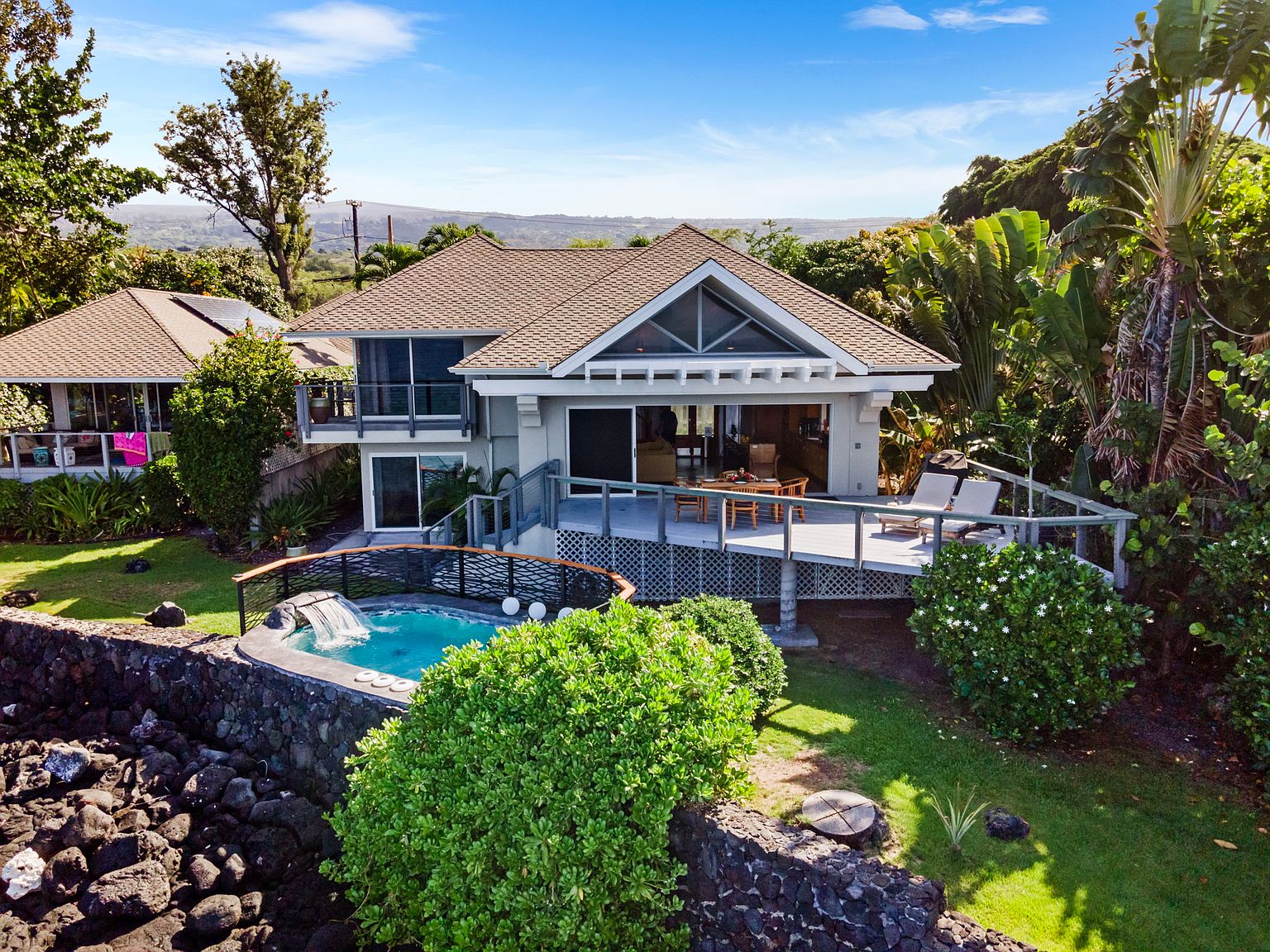 This aerial view showcases a charming coastal home with a well-maintained yard and a unique spa. The house features a deck with outdoor seating, perfect for enjoying the ocean breeze. The spa, nestled against a rock wall, adds a touch of luxury and privacy to the property.