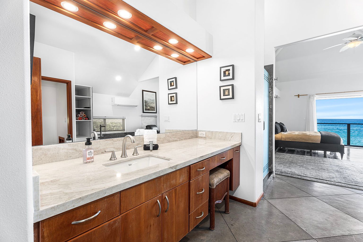 This is a well-lit primary bathroom featuring a wooden vanity with a marble countertop and a rectangular sink. A large mirror with overhead lighting enhances the space, while framed artwork adds a touch of elegance. The bathroom opens into a bedroom with an ocean view, creating a luxurious and relaxing atmosphere.