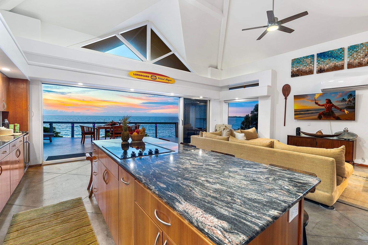 This interior shot showcases a modern kitchen that seamlessly blends indoor and outdoor living. The kitchen features a large island with a striking countertop, wooden cabinetry, and an open layout that flows into the living area. Sliding glass doors reveal a stunning ocean view and access to an outdoor deck, creating a luxurious and inviting atmosphere.