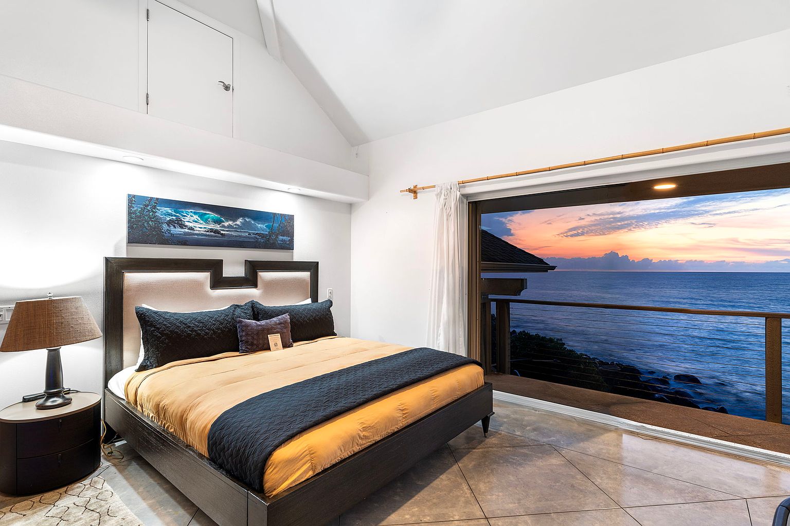 This primary bedroom features a king-sized bed with a dark wood frame, complemented by a beige headboard and a mix of dark and textured pillows. A large sliding glass door opens to a balcony with ocean views, enhancing the room's appeal. The room is well-lit, showcasing a blend of modern design and serene coastal living.