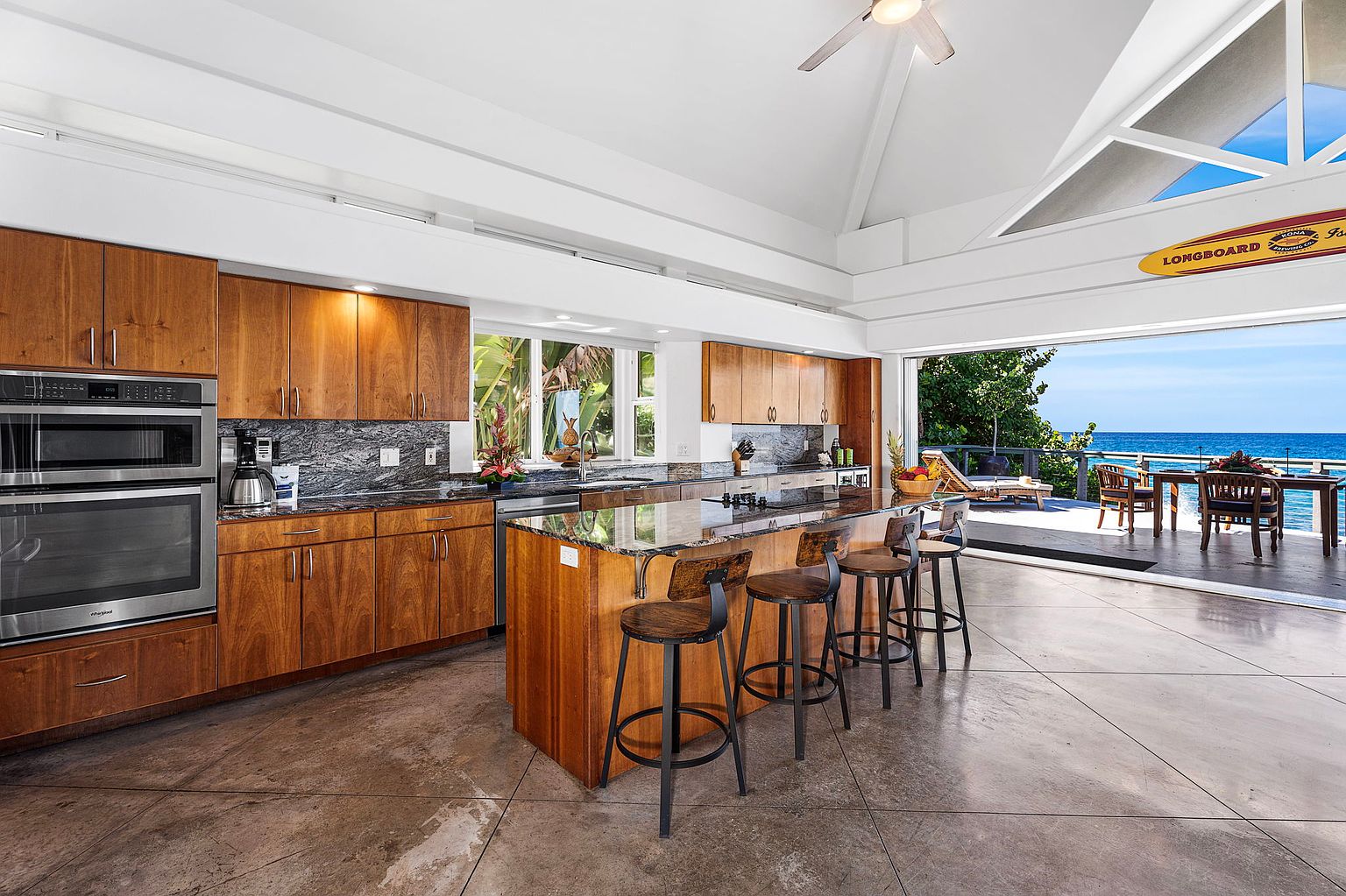 This is a bright and airy kitchen with wooden cabinetry and a large island with bar seating. The kitchen opens up to an outdoor patio/deck area with ocean views, creating a seamless indoor-outdoor living experience. Stainless steel appliances and granite countertops add a touch of luxury.