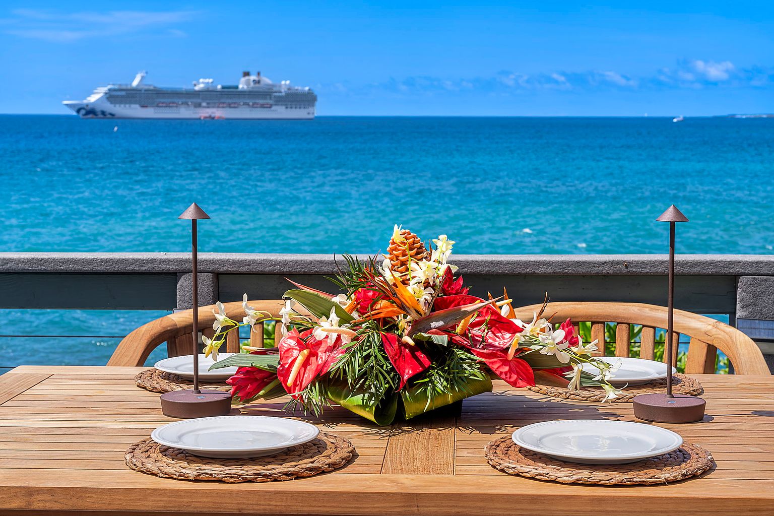 This image showcases a beautifully set outdoor dining area on a patio or deck, featuring a wooden table adorned with a vibrant floral centerpiece, place settings, and stylish lamps. The backdrop offers a stunning ocean view with a cruise ship in the distance, creating a luxurious and inviting atmosphere. The scene suggests a perfect setting for outdoor entertaining and relaxation.