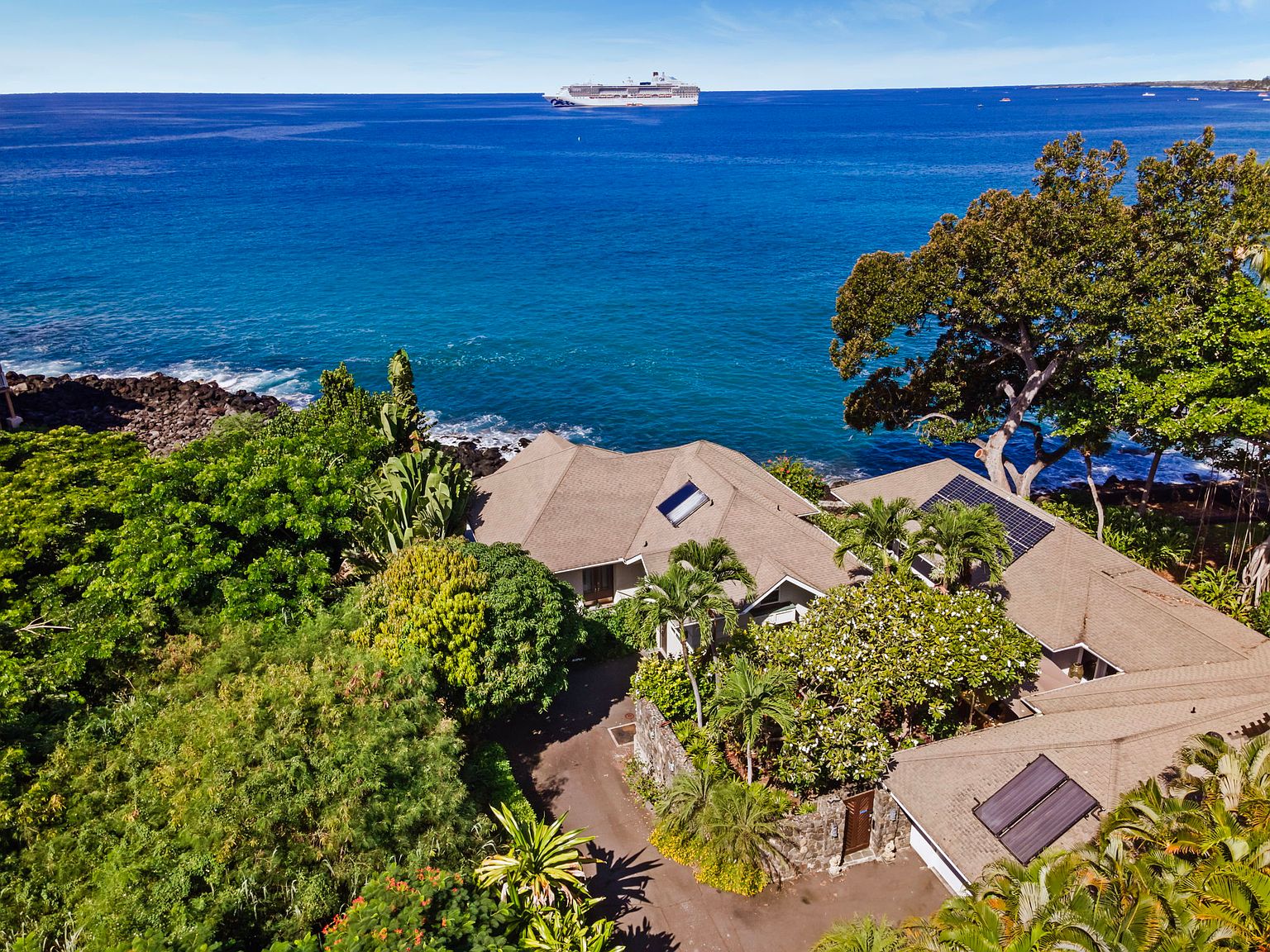 This aerial shot showcases a luxurious coastal property with a sprawling house nestled amidst lush greenery. The house features a complex roofline with solar panels and a skylight, suggesting modern amenities. The property overlooks the ocean, with a large ship visible in the distance, emphasizing the prime waterfront location.