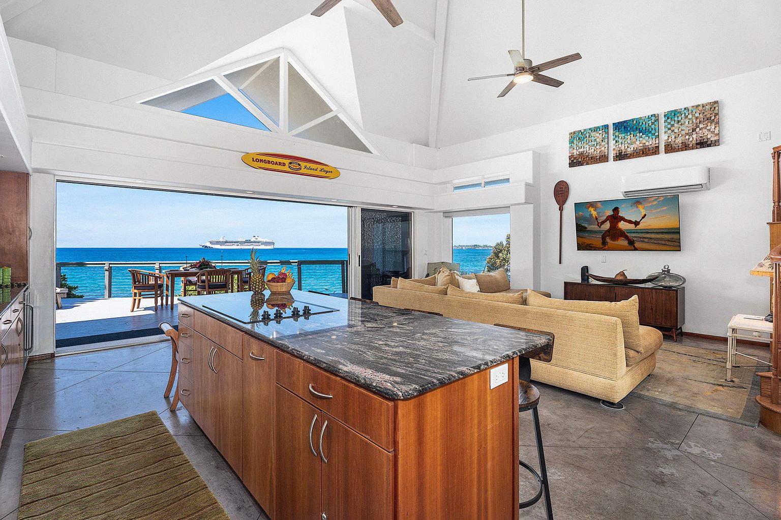 This interior shot showcases a bright and airy living space with an open floor plan, seamlessly blending indoor and outdoor living. The kitchen island with a dark countertop is in the foreground, leading to a living area with a tan sofa and a view of the ocean through large sliding glass doors. The room features modern decor, including artwork and a flat-screen TV, creating a luxurious and inviting atmosphere.