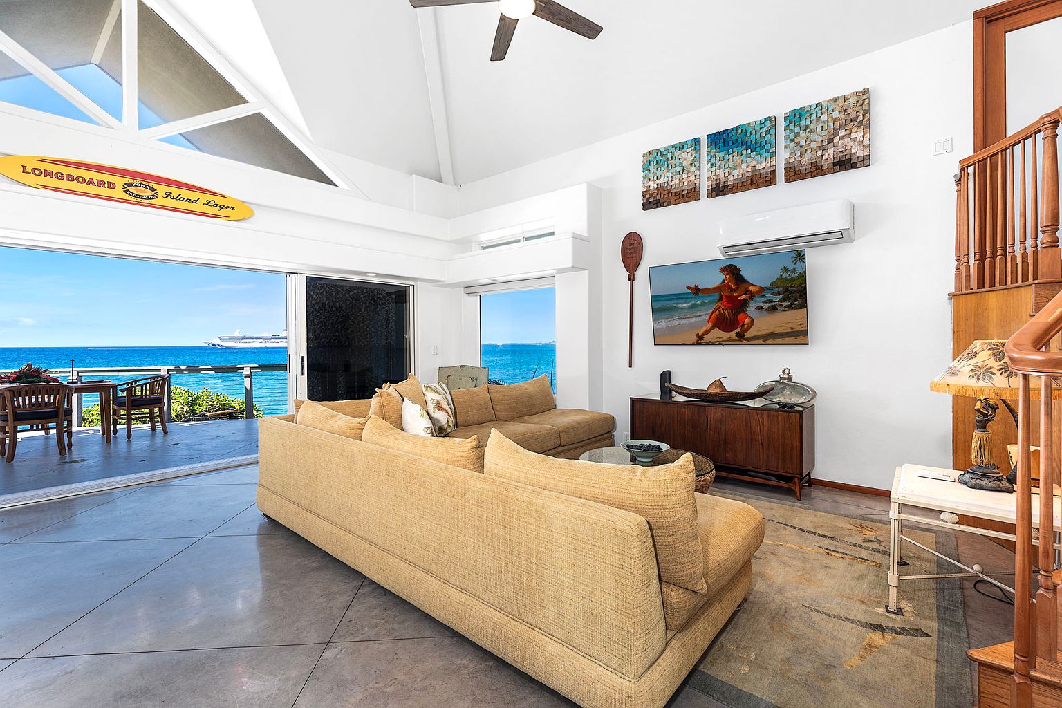 This is an interior shot of a living room with an open floor plan that leads to an outdoor patio overlooking the ocean. The room features a large, comfortable-looking sectional sofa, a wooden entertainment center with a television, and artwork on the walls. The style is coastal and relaxed, with natural light flooding the space, creating a welcoming and luxurious atmosphere.