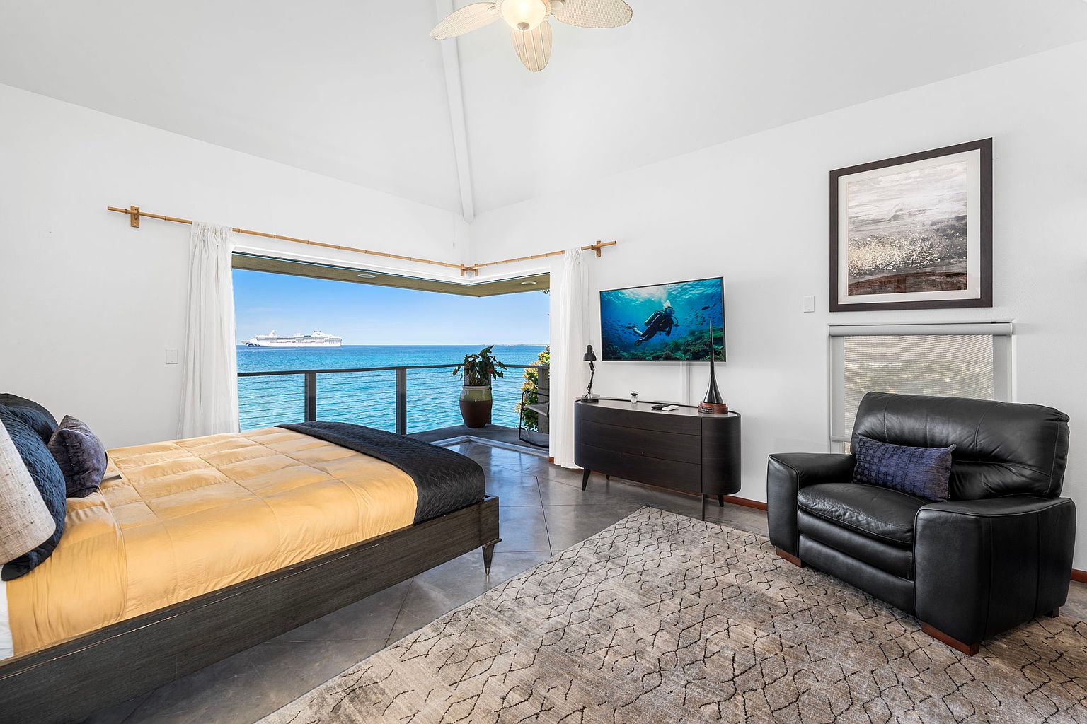 This primary bedroom features a bed with a yellow comforter, a dark wood frame, and blue pillows. A large sliding glass door opens to a balcony with ocean views. The room also includes a dark wood dresser with a television, a black leather armchair, and a patterned area rug, creating a luxurious and relaxing atmosphere.