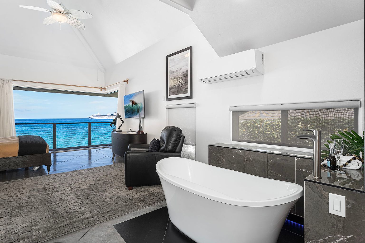 This is a luxurious primary bathroom featuring a freestanding white bathtub as the focal point, complemented by dark marble countertops and cabinetry. The room boasts an ocean view through a large window, enhancing the spa-like atmosphere. A modern black leather chair and a gray rug add a touch of comfort and style to the space.