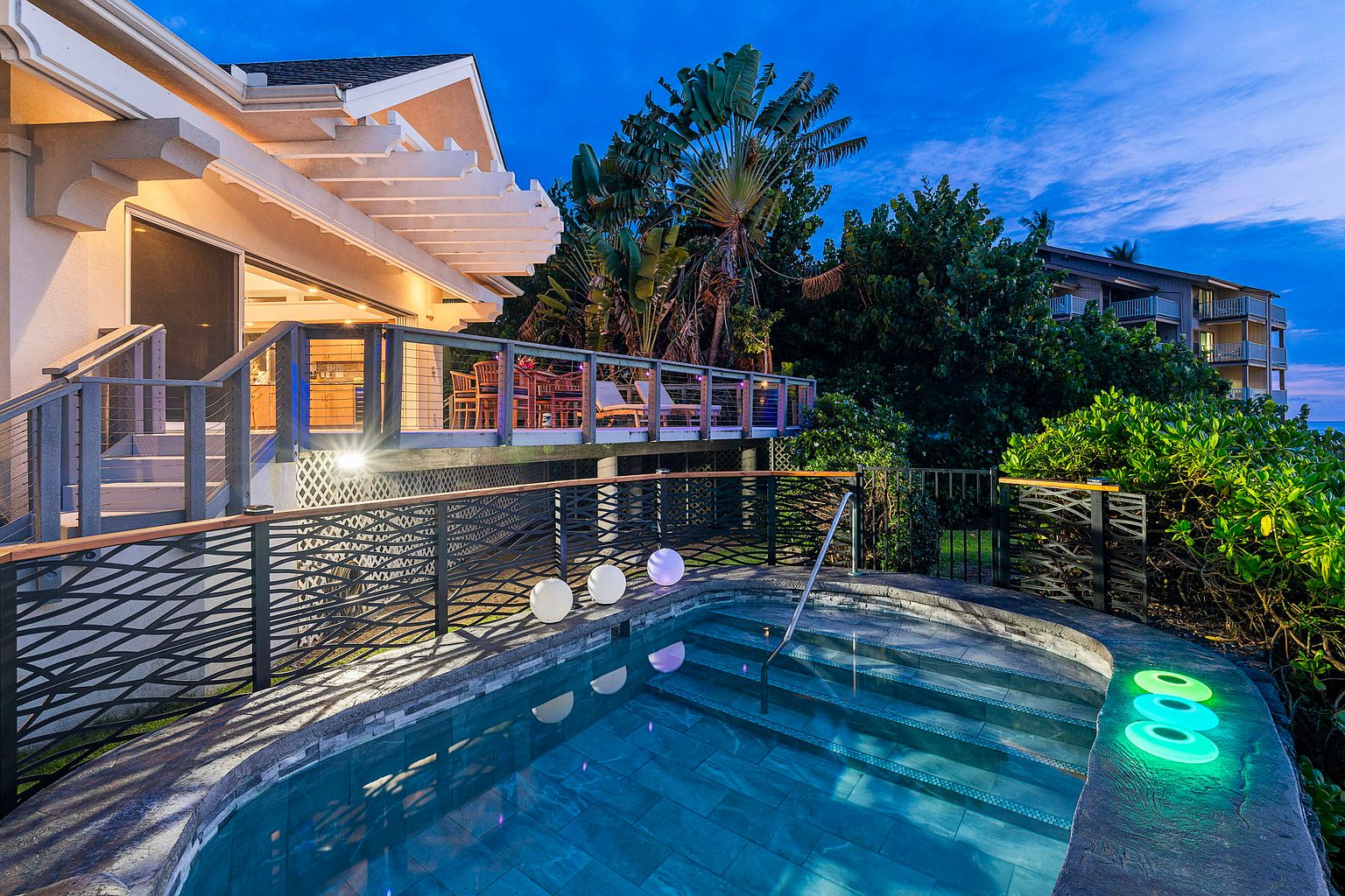 This image showcases a luxurious outdoor spa area at dusk, featuring a small pool with illuminated steps and decorative lighting. The pool is surrounded by a stone patio and a modern black metal fence with a wooden top rail. A raised deck with outdoor furniture is visible in the background, suggesting an inviting space for relaxation and entertainment.