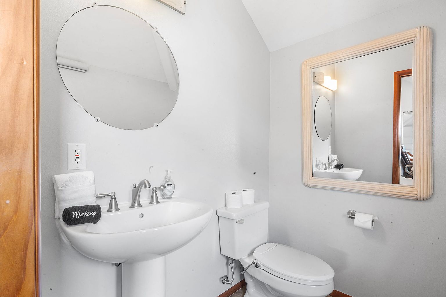 This is a well-lit guest bathroom featuring a pedestal sink with a round mirror above it, complemented by a square mirror on the adjacent wall. A toilet with a bidet attachment is positioned next to the sink, and the walls are painted in a neutral tone. The overall impression is clean and functional.