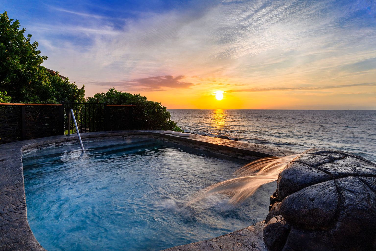 This image showcases a luxurious spa or pool overlooking the ocean at sunset. The pool features a unique rock waterfall feature, adding a natural and serene element. The scene evokes a sense of relaxation and high-end living, perfect for attracting potential buyers seeking a tranquil retreat.