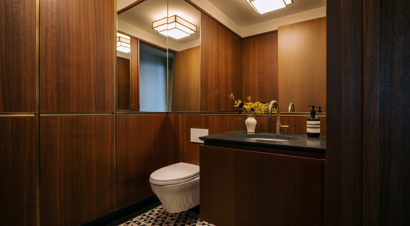 This sophisticated powder room features rich, warm wood-paneled walls and a striking geometric patterned floor. The space is accented by a modern brass faucet, a sleek dark stone vanity, and a large mirror that enhances the room's depth. A contemporary square light fixture provides a warm, inviting glow, creating a luxurious and polished atmosphere.