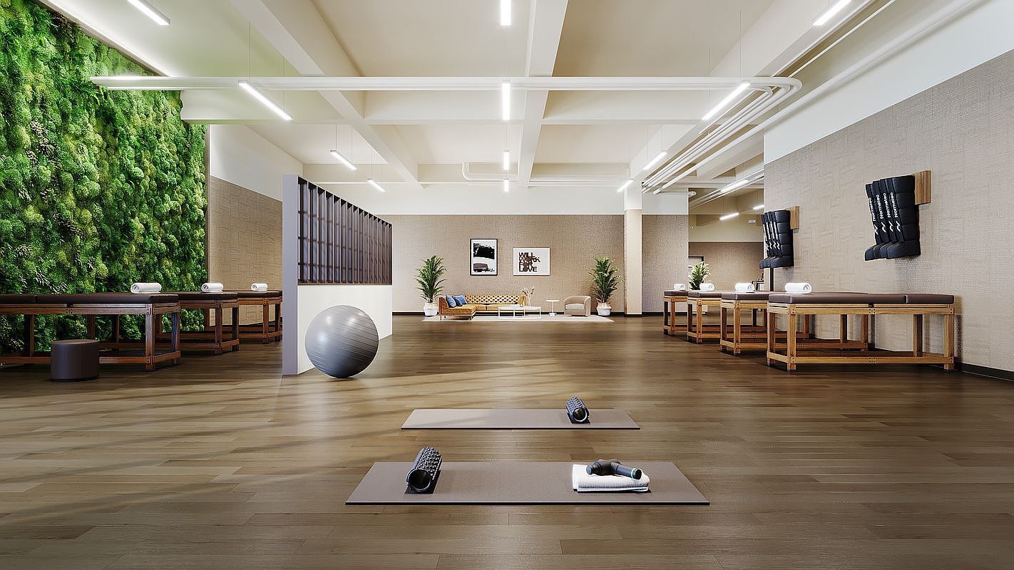 This spacious, modern wellness studio features a striking living moss wall on the left and multiple massage tables arranged for recovery sessions. The room is anchored by warm wood flooring, minimalist decor, and a central lounge area, creating a serene and professional atmosphere. The wide-angle perspective emphasizes the open layout, highlighting the integration of fitness and relaxation amenities.
