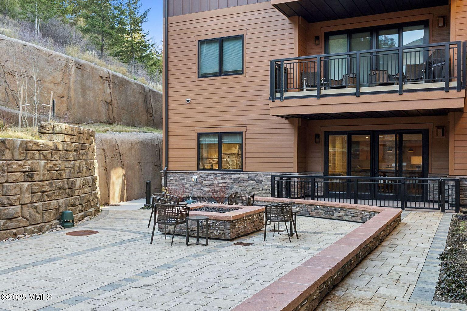 This image showcases a beautifully designed outdoor patio area. The patio features a stone fire pit surrounded by comfortable seating, creating an inviting space for relaxation and entertainment. The area is paved with patterned stones and bordered by low stone walls, complemented by the building's exterior with its brown siding and black-framed windows and railings. The patio is nestled against a natural rock wall, adding a unique and appealing backdrop.