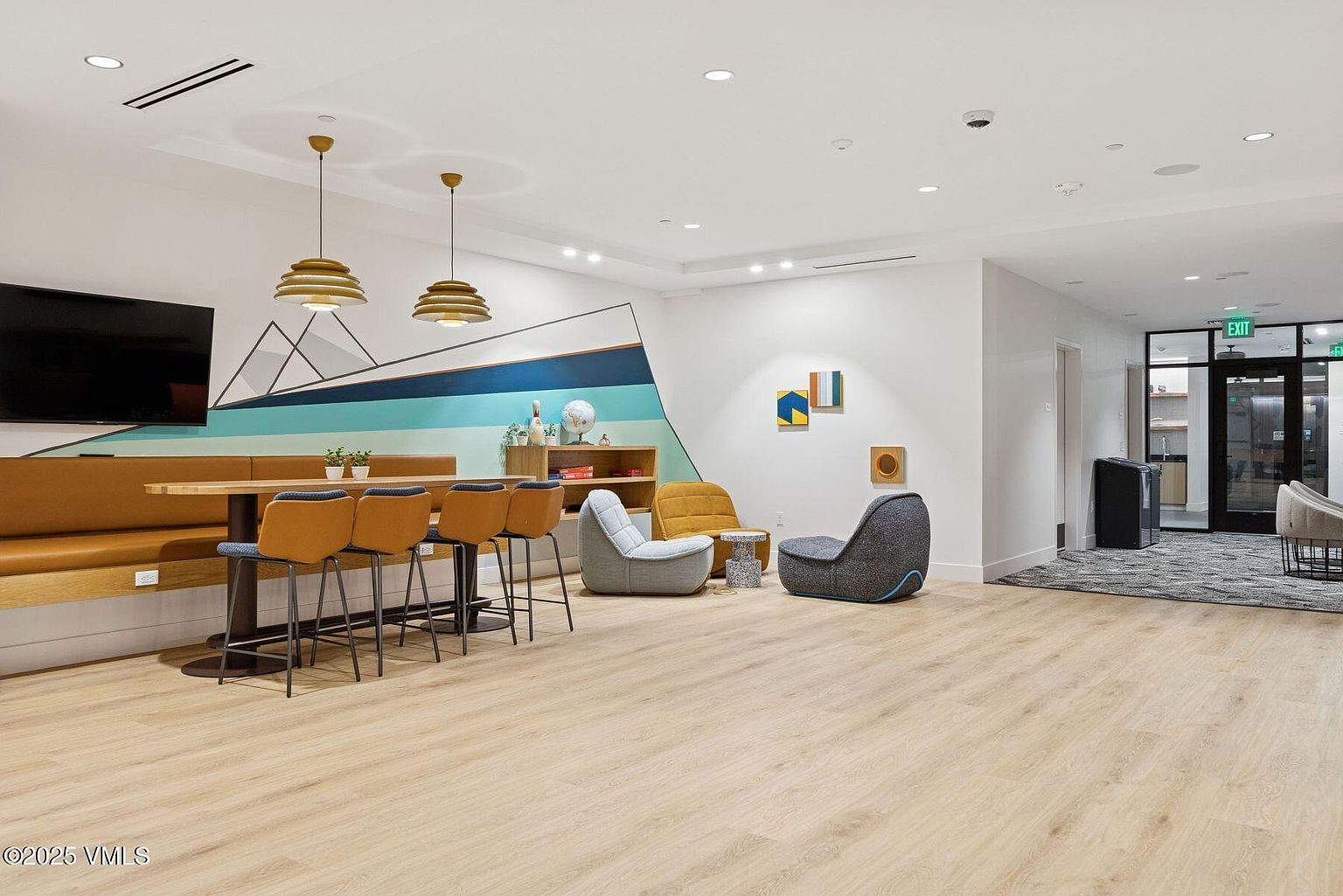 This is an interior shot of a community amenity space, featuring a long table with built-in seating and bar stools, complemented by lounge chairs and modern artwork. The space is well-lit with a combination of recessed lighting and pendant fixtures, creating a welcoming and contemporary atmosphere. The flooring is light-colored wood, and the overall impression is clean and inviting.