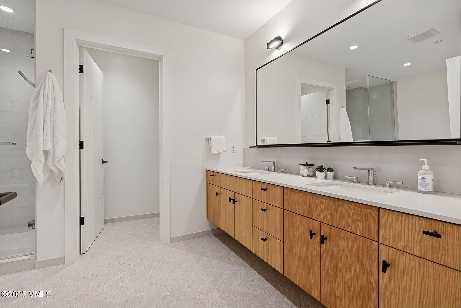 This is a bright and modern primary bathroom featuring a double vanity with light wood cabinetry and black hardware. A large, rectangular mirror with a black frame hangs above the vanity, reflecting the clean lines of the space. The flooring is a light-colored herringbone pattern, and a glass-enclosed shower is visible to the left, contributing to the bathroom's spa-like atmosphere.