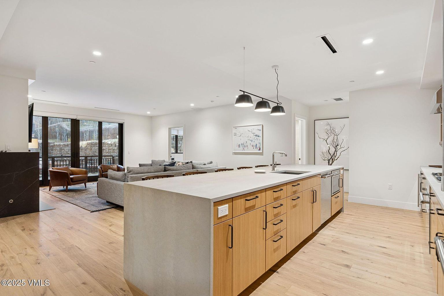 This is a bright and modern kitchen that seamlessly blends into the living space. The kitchen features a large island with a concrete countertop and wooden cabinetry, stainless steel appliances, and pendant lighting. The open floor plan allows for easy flow between the kitchen, dining, and living areas, creating an inviting and functional space.