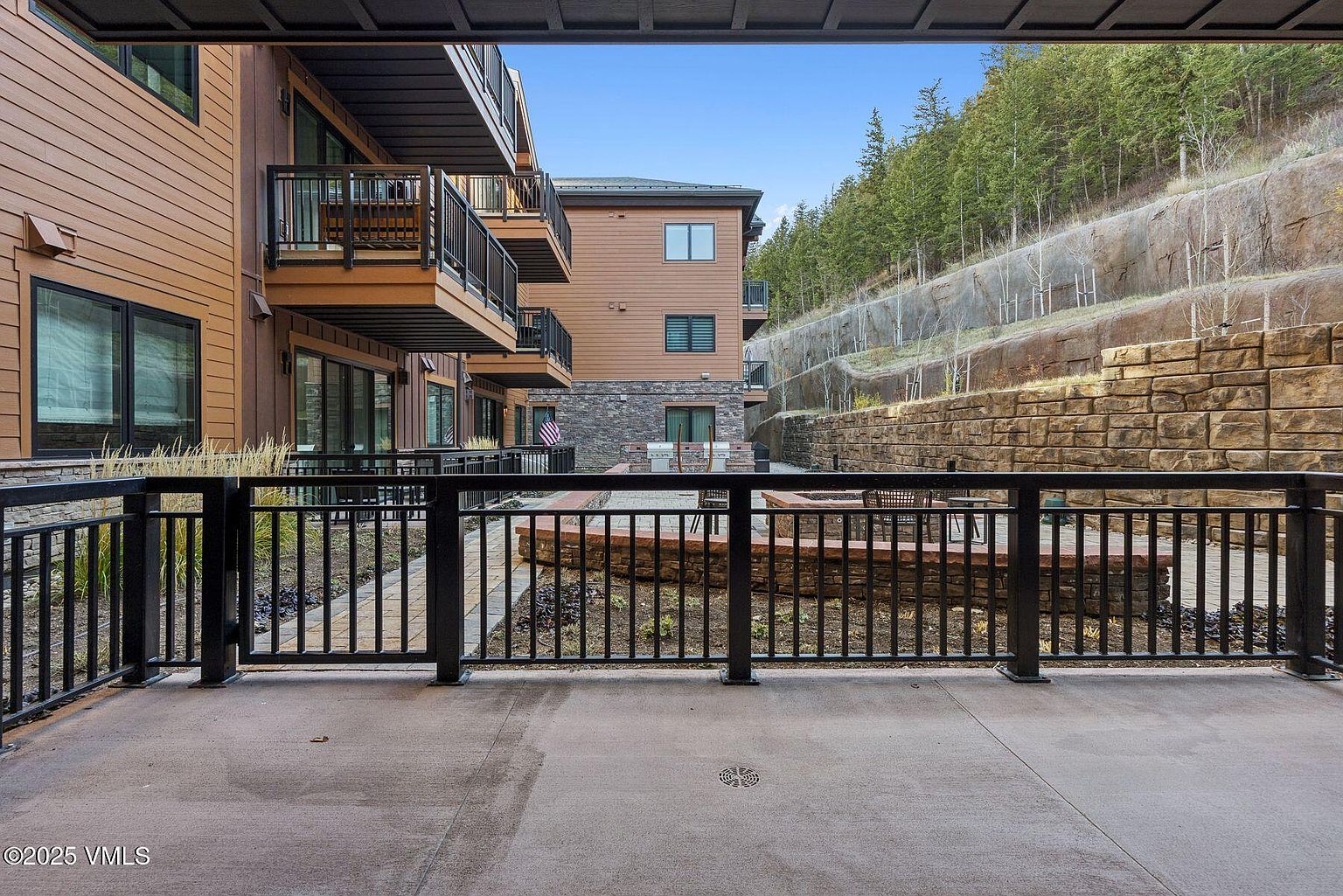 This image showcases a well-maintained patio or balcony area, featuring a black metal railing that provides a clear view of the outdoor space. The patio is constructed of concrete and overlooks a landscaped area with a brick retaining wall and a fire pit. The backdrop includes a building with wood siding and balconies, as well as a hillside with trees, creating a serene and inviting outdoor living space.