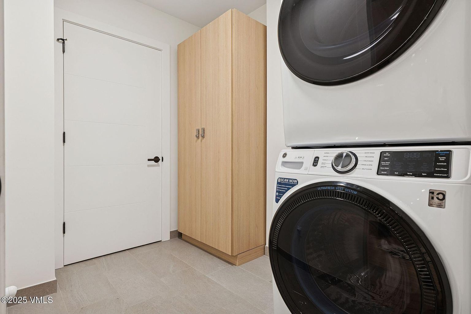 This is an interior shot of a laundry room featuring a stacked washer and dryer set in the foreground. A light wood-colored storage cabinet and a white door with modern hardware are visible in the background. The flooring appears to be light-colored tile, contributing to a clean and functional space.