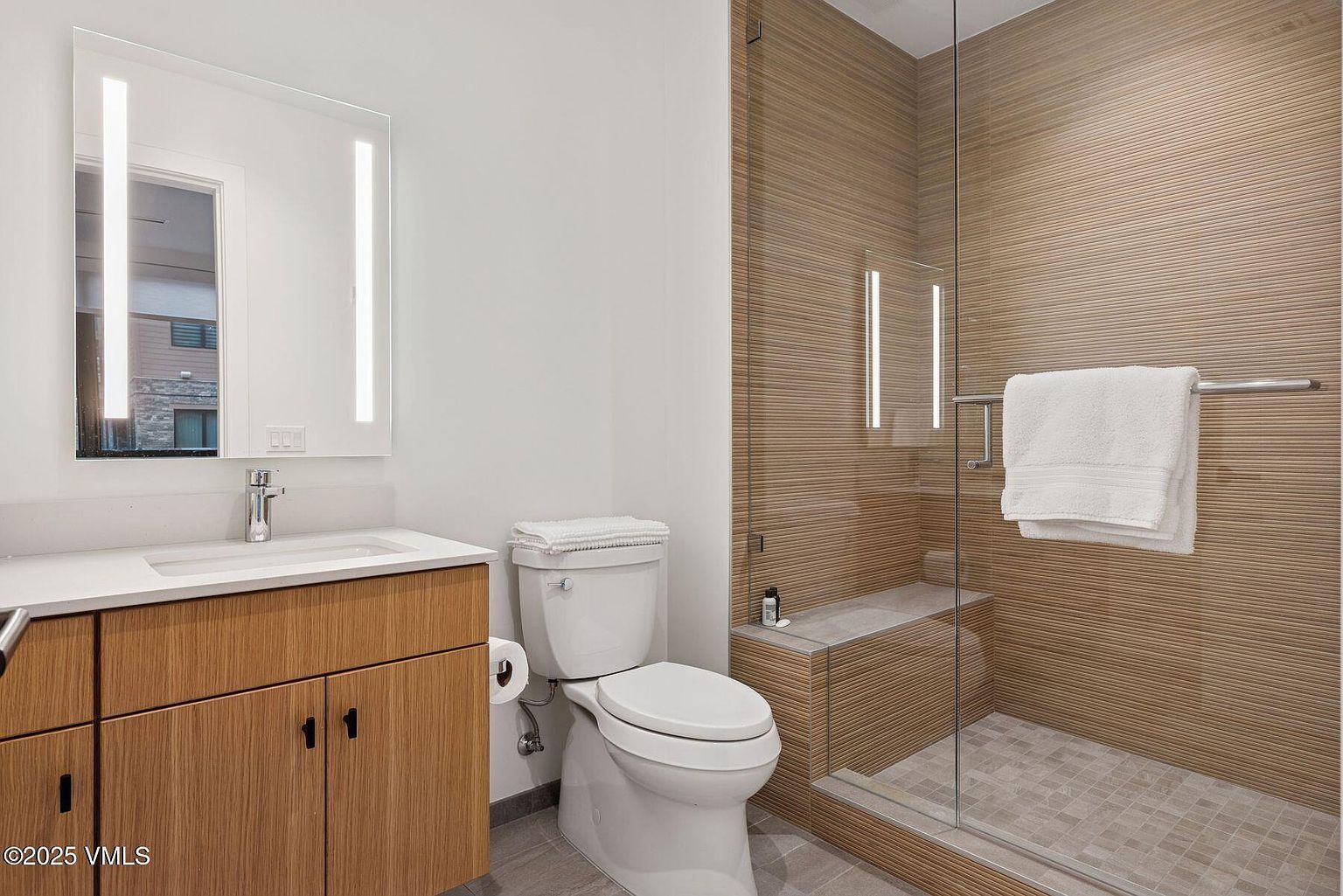 This is a well-lit bathroom featuring a modern design with a wood-paneled vanity, a white countertop, and a sleek mirror with integrated lighting. A toilet with a white tank and bowl sits next to the vanity, and a glass-enclosed shower with matching wood-paneled walls and a built-in bench is visible. The perspective is a wide shot, capturing the entire bathroom space.