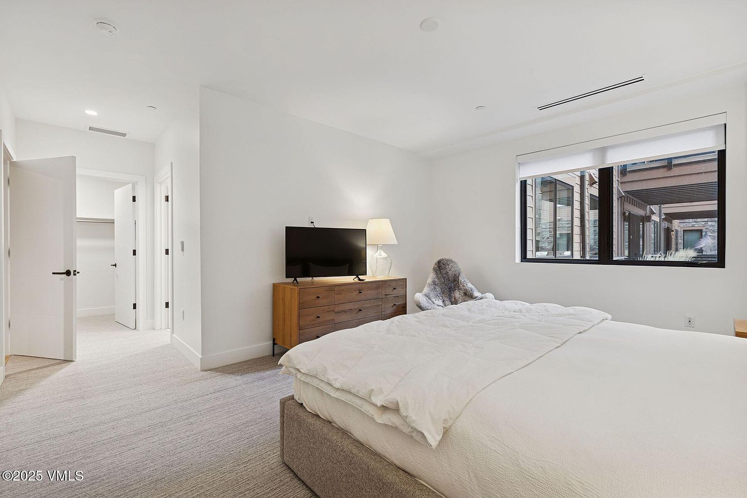 This is a bright and airy bedroom featuring a neutral color palette. The room includes a bed with white linens, a wooden dresser with a television, and a window providing natural light. A doorway leads to a hallway, and the overall impression is clean and modern.