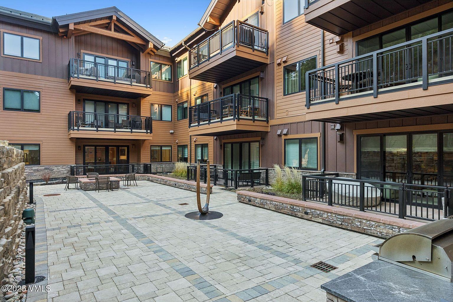 This image showcases a beautifully designed outdoor courtyard area of a multi-story residential building. The courtyard features a paved patio with seating arrangements, complemented by well-maintained landscaping and balconies overlooking the space. The architectural style is modern with wood and stone accents, creating an inviting and upscale atmosphere.