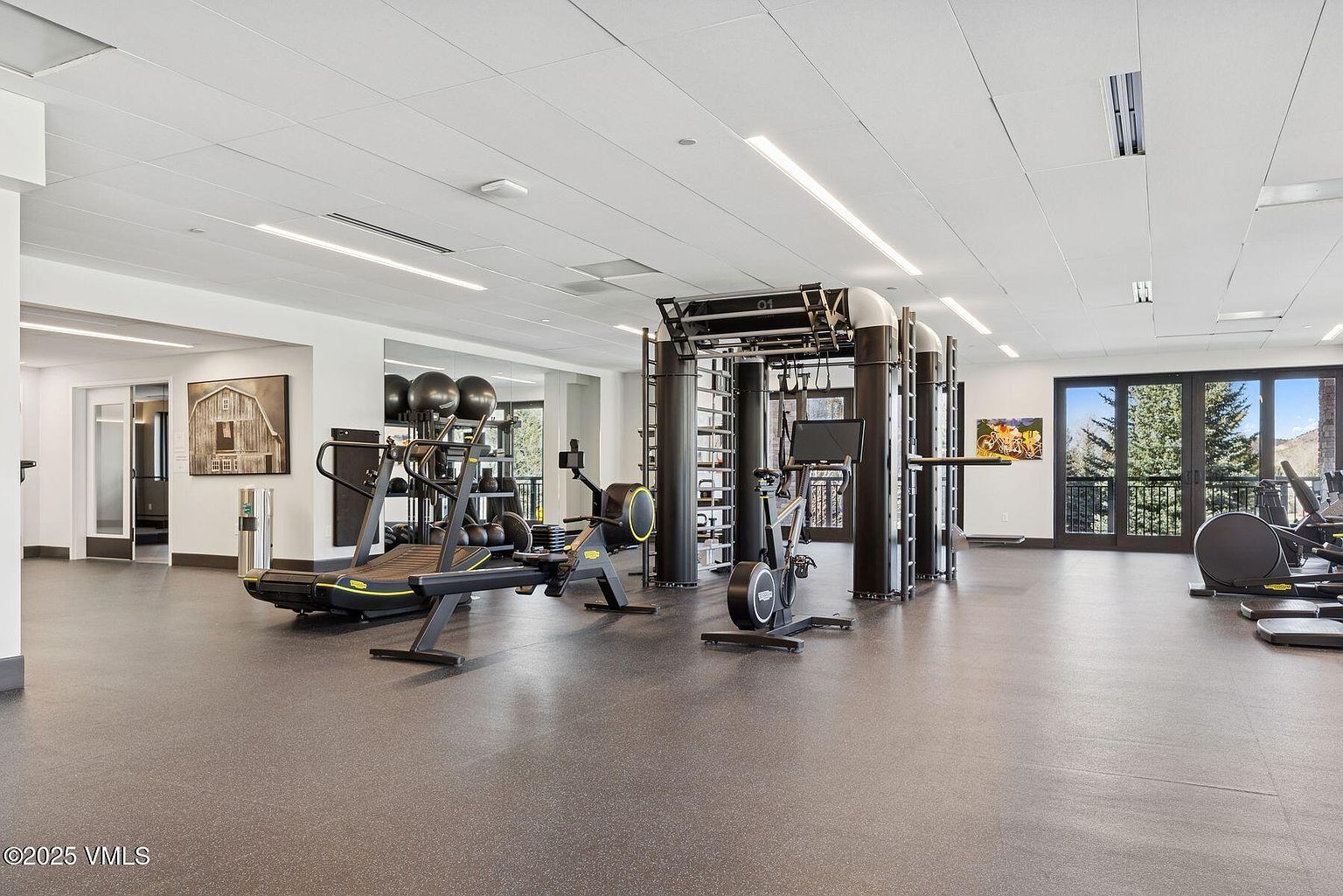 This is a spacious and well-equipped home gym featuring various exercise machines such as treadmills, rowing machines, and stationary bikes. The room has a neutral color palette with gray flooring and white walls, creating a clean and modern aesthetic. Large windows provide natural light and views of the outdoors, enhancing the workout experience.