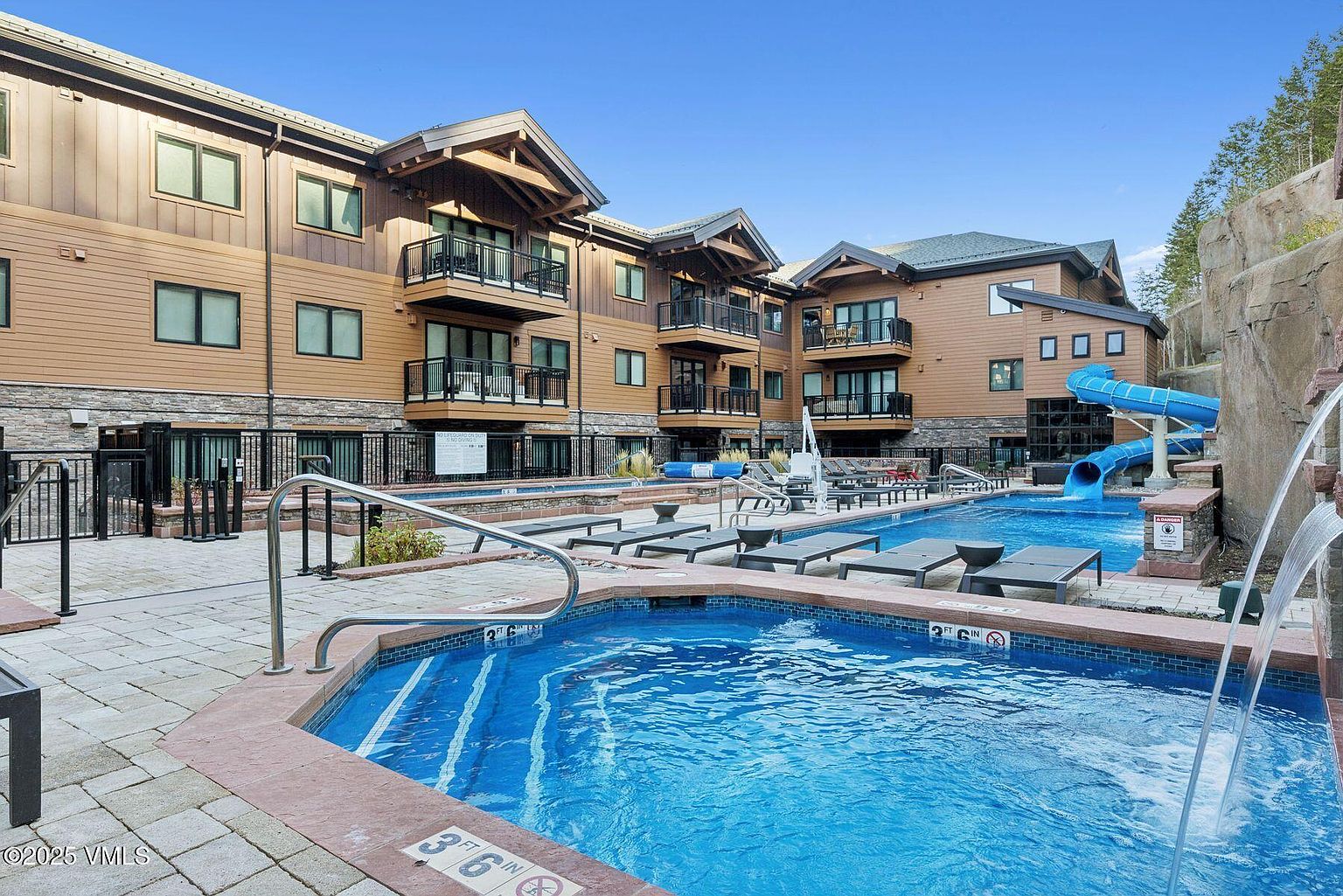 This image showcases a luxurious outdoor pool and spa area of a multi-story building. The pool features a water slide and lounge chairs, while the spa is a raised hot tub with clear blue water. The building has a modern architectural style with wood and stone accents, and balconies overlooking the pool area.