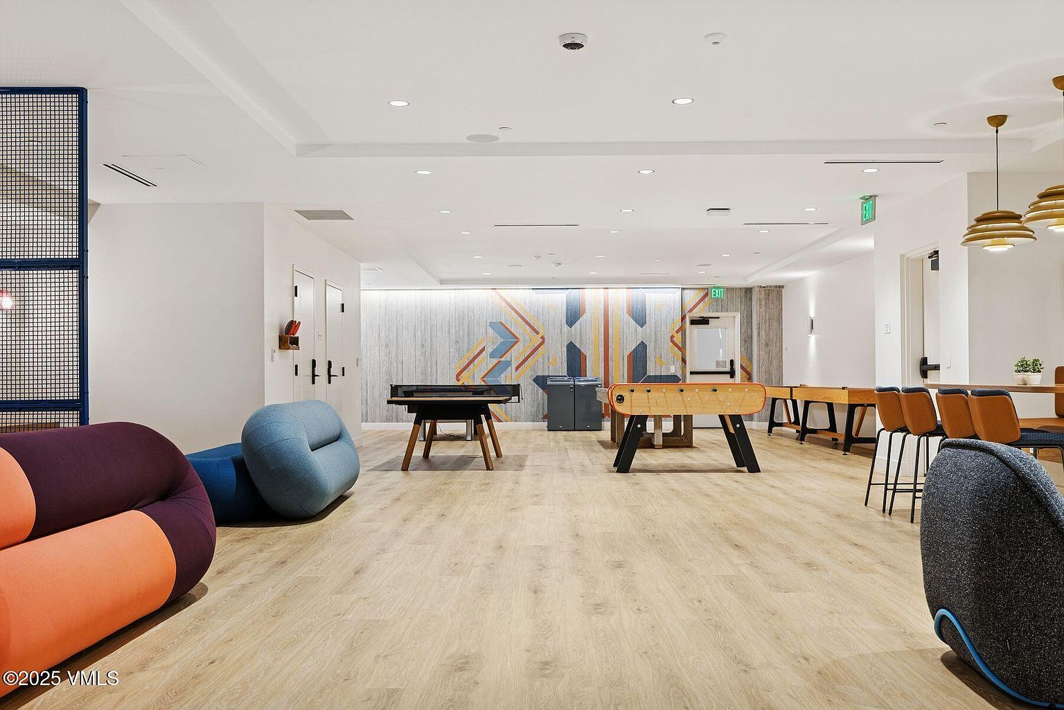 This is an interior shot of a community amenity space, featuring a foosball table, ping pong table, and comfortable lounge seating. The space is well-lit with a modern design, including light wood flooring and a geometric accent wall. The room appears to be a recreational area designed for residents to socialize and enjoy leisure activities.