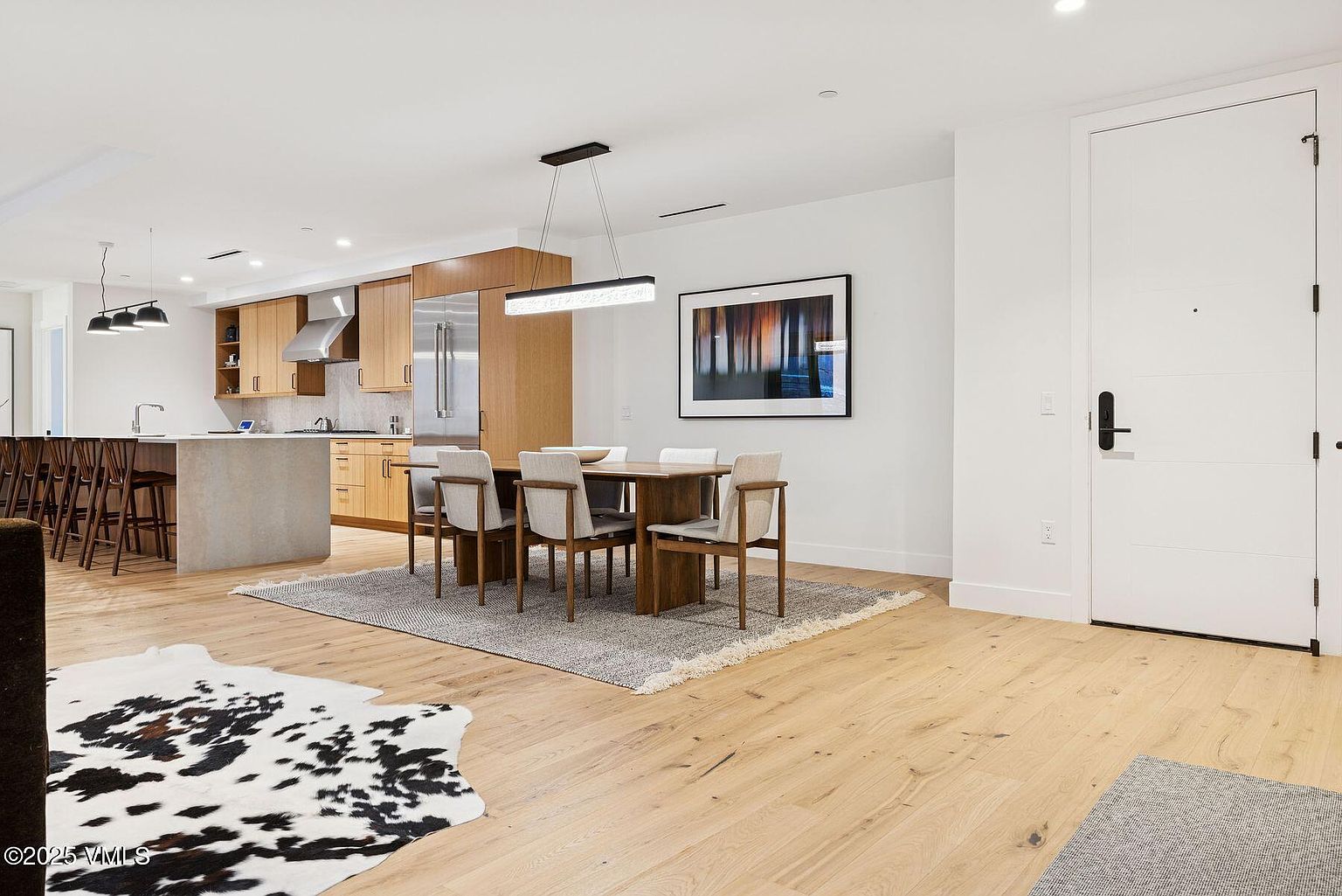 This interior shot showcases a modern dining area adjacent to a kitchen. The dining set includes a wooden table and six chairs, positioned on a gray rug with fringed edges. The room features light hardwood flooring, white walls, and a contemporary light fixture hanging above the table, creating a bright and inviting space.